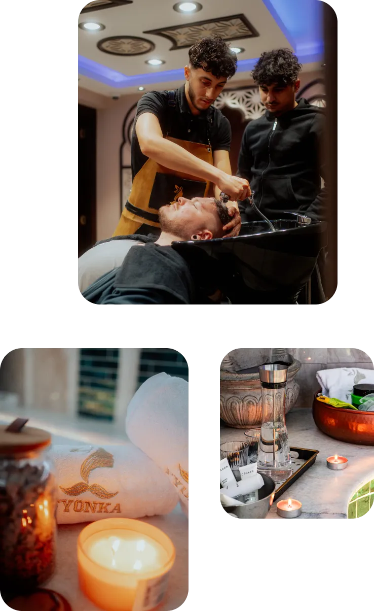 Ein Friseur wäscht bei einem Kunden die Haare, während eine weitere Person zusieht, neben einer Einstellung mit einer brennenden Kerze und einem Turbanhandtuch sowie einem Tablett mit Wasserkaraffe, Gläsern und Teelichtern auf einem Marmorwaschtisch.