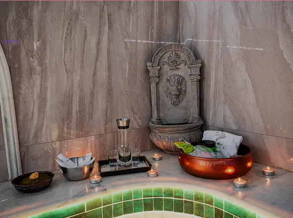 Stilvoller Spa-Bereich mit Marmorfliesen, brennenden Teelichtern, einer dekorativen Wandbrunnen-Skulptur mit Löwenkopf und mit Wasser gefüllten Schalen.