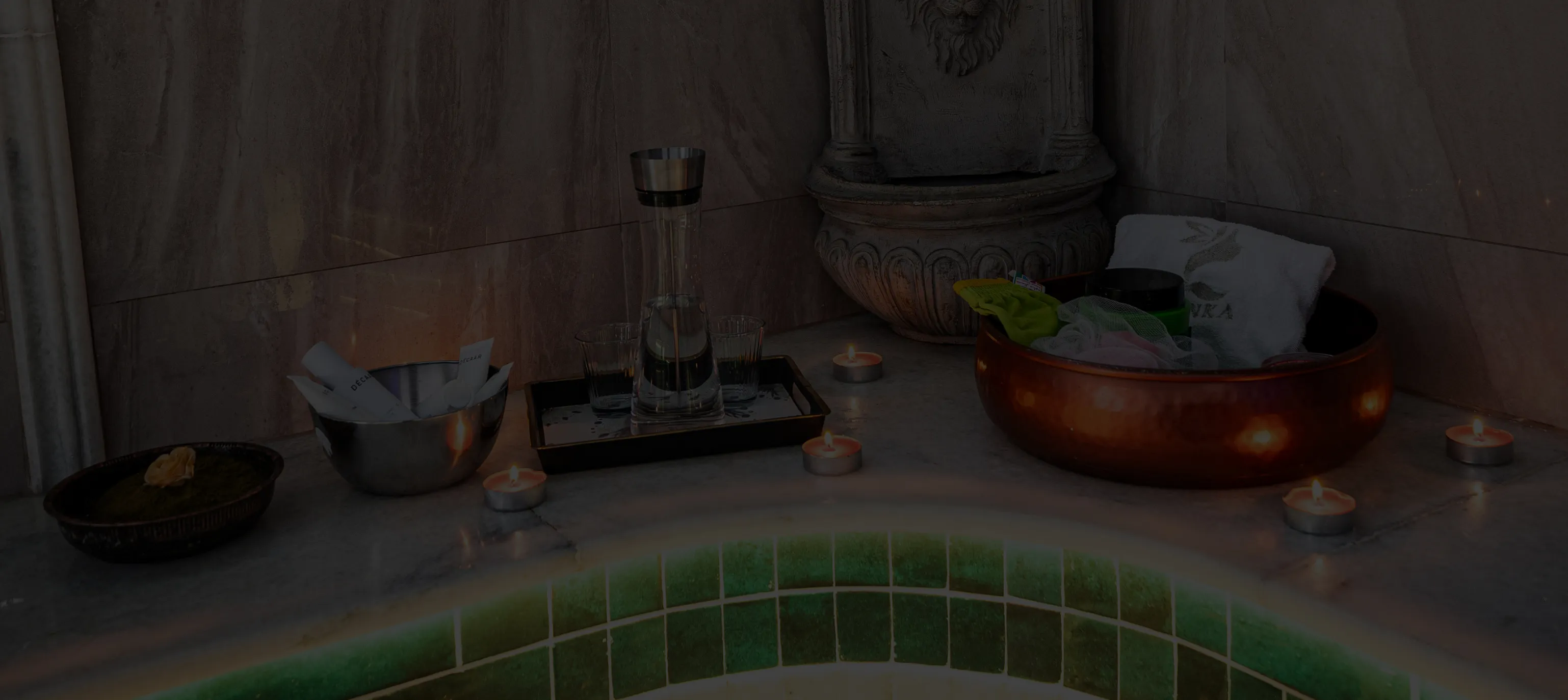 Wellnessbereich mit brennenden Teelichtern, Wasserkrug mit Gläsern und Schale mit Handtüchern auf einem Marmortisch.