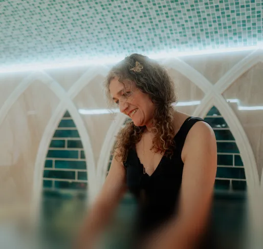 Lächelnde Frau mit lockigem Haar und Schmetterlings-Haarklammer vor einer hellen Wand mit Mosaikfliesen.