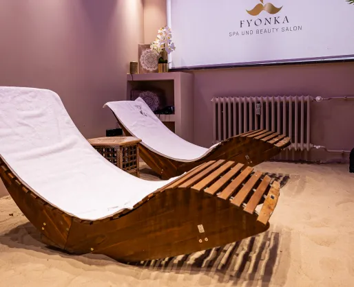 Zwei Holzliegen mit weißen Polstern in einem ruhigen Spa-Raum mit Sandboden und Dekoration.