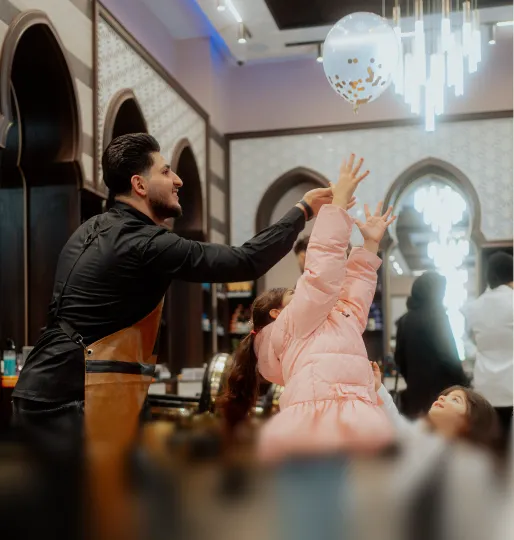 Mann in schwarzem Hemd und brauner Schürze wirft einen Ballon mit Goldkonfetti, während zwei Kinder im Innenraum eines Restaurants danach greifen.