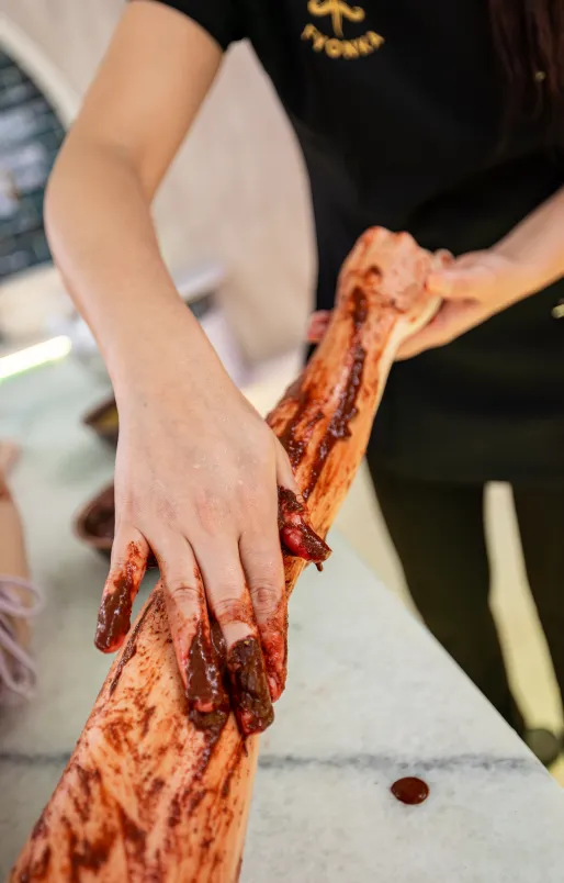 Eine Person trägt eine Schürze und bedeckt die Arme einer anderen Person mit geschmolzener Schokolade.