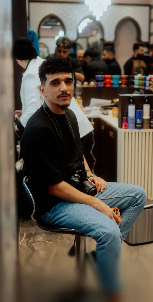 Junger Mann sitzt mit Kamera um den Hals und Handy in der Hand in einem belebten Friseursalon.