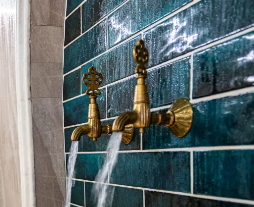 Zwei laufende goldene Wasserhähne an einer Wand mit grünen Fliesen.