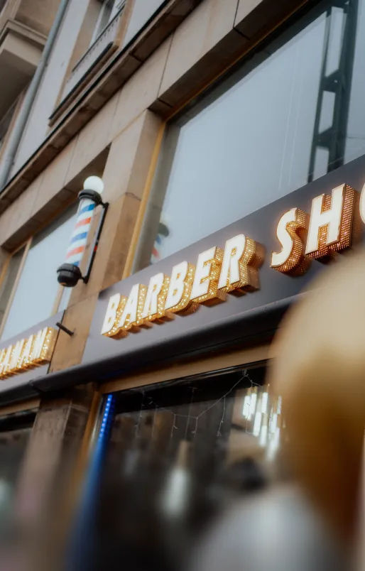 Außenansicht eines Friseurladens mit beleuchtetem BARBER SHOP Schild und klassischem rot-weiß-blauem Barbier-Polssteuer.