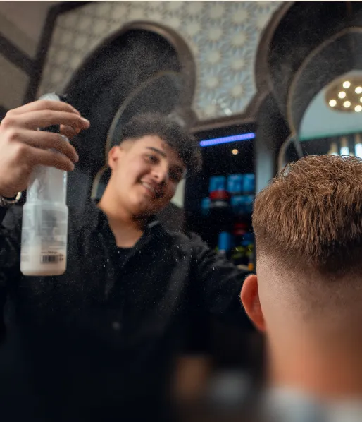 Friseur sprüht Haarspray auf einen jungen Mann mit kurzem, gestyltem Haar im Friseursalon.
