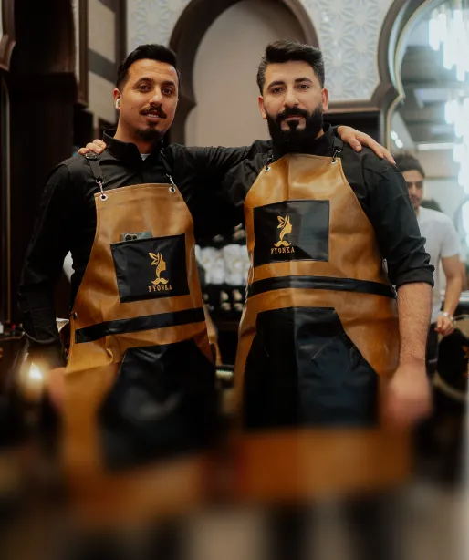 Zwei Männer in braunen ledernen Schürzen mit dem Logo Fyona, die Arm in Arm in einem Friseursalon stehen.