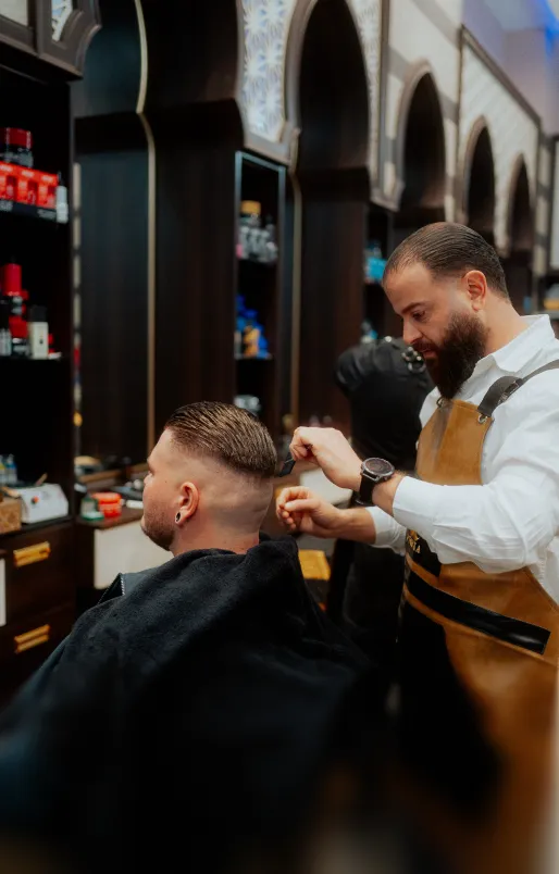 Friseur mit braunem Bart schneidet einem Mann in einem Friseursalon mit dunklen Holzregalen und arabischem Design die Haare.