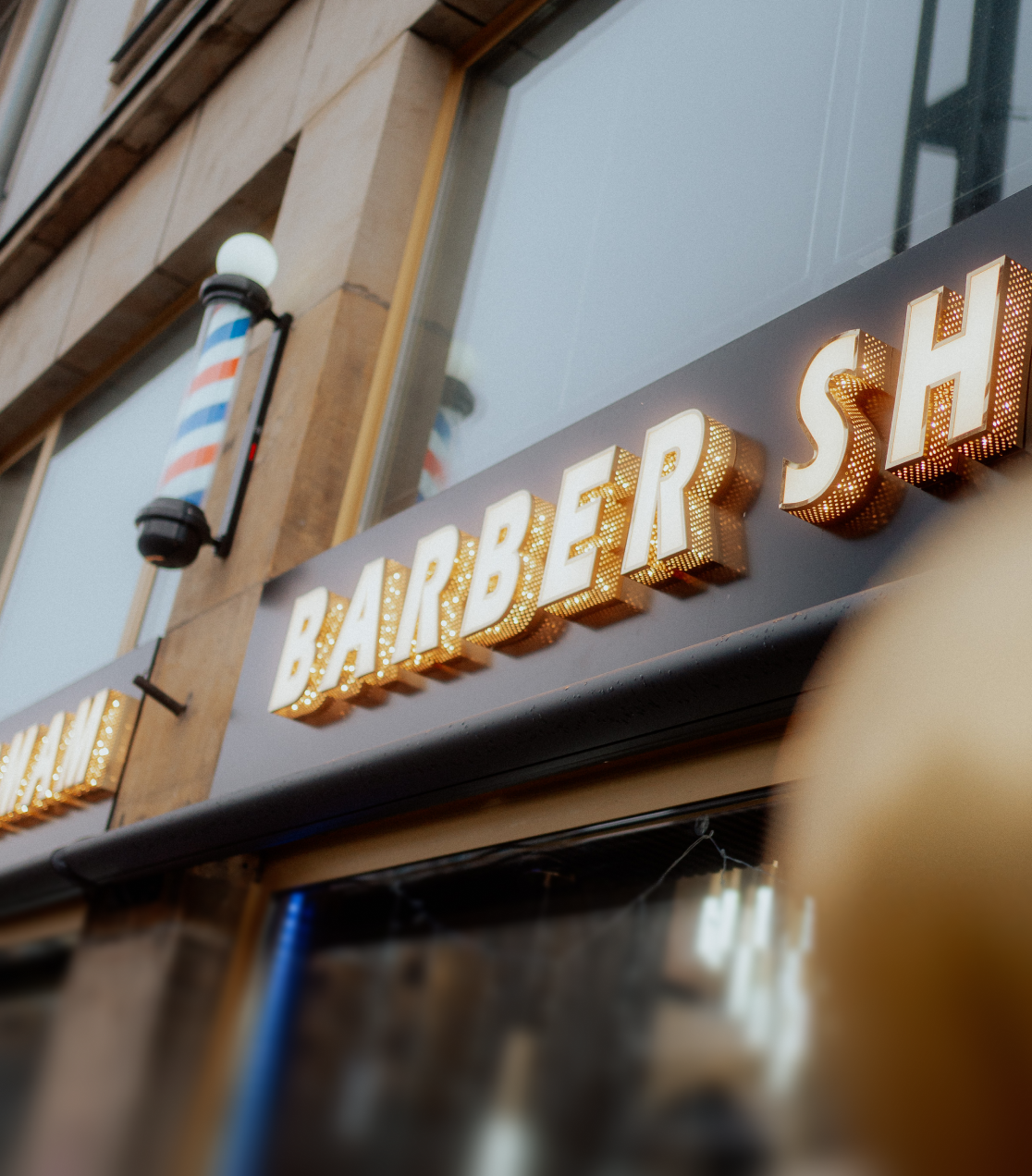 Leuchtreklame eines Friseursalons mit beleuchteten Buchstaben und einem traditionellen rot-blau-weißen Barbierstab an der Außenwand.