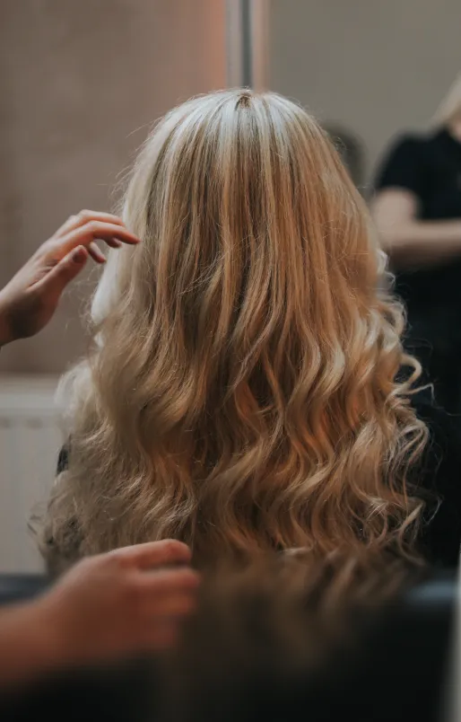 Rückansicht einer Person mit langen, welligen blonden Haaren, die von einer anderen Person frisiert wird.
