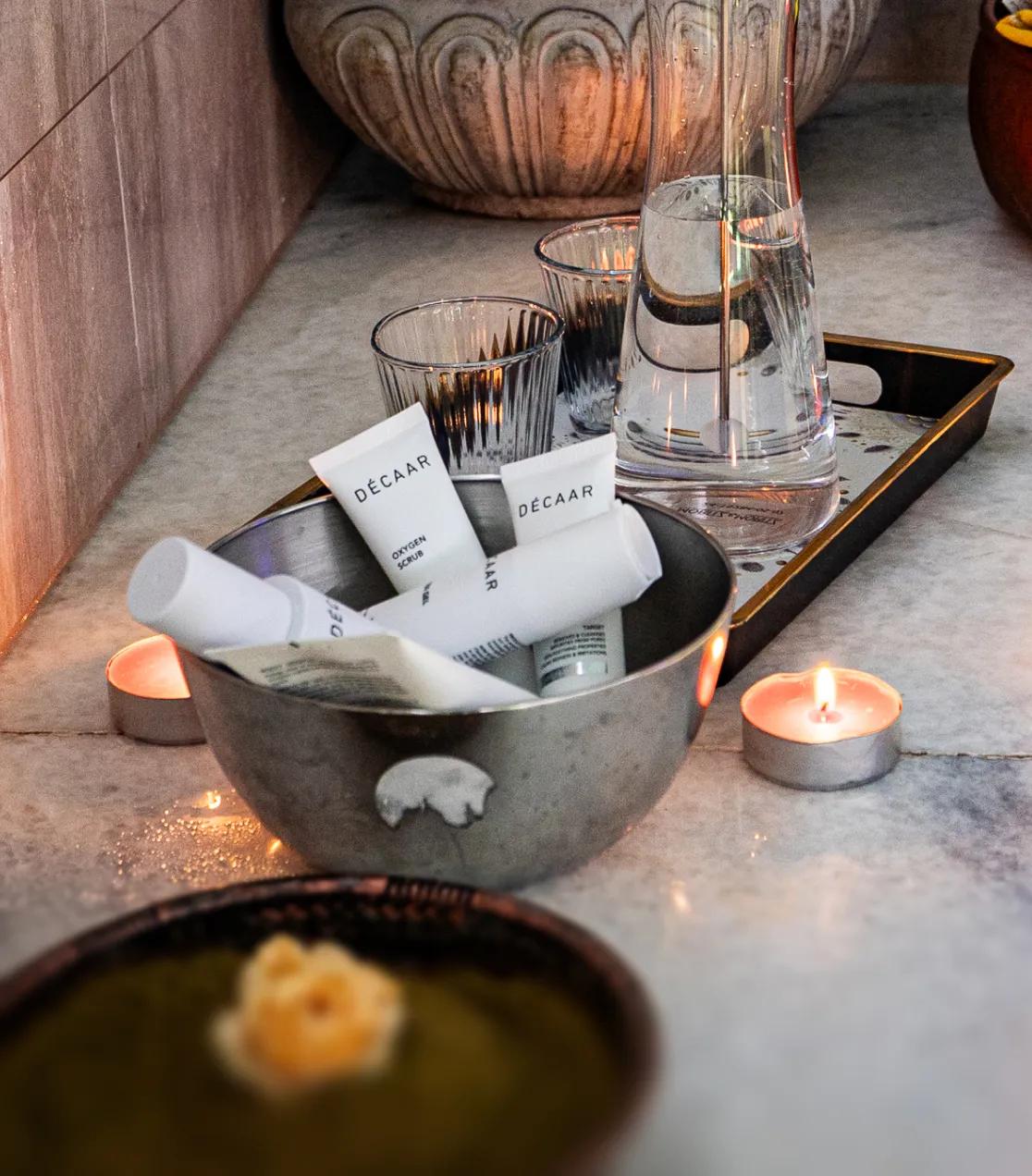 Wellness-Arrangement mit DÉCAAR Hautpflegeprodukten in einer Metallschale, zwei Wassergläsern, einer Karaffe und zwei brennenden Teelichtern auf Marmorfläche.