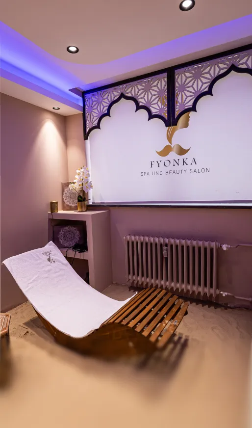 Entspannender Spa-Raum mit ergonomischem Holzliegestuhl, weißen Kissen, Orchideen und einem beleuchteten Veluna Wellness House Spa und Beauty Salon-Logo an der Wand.