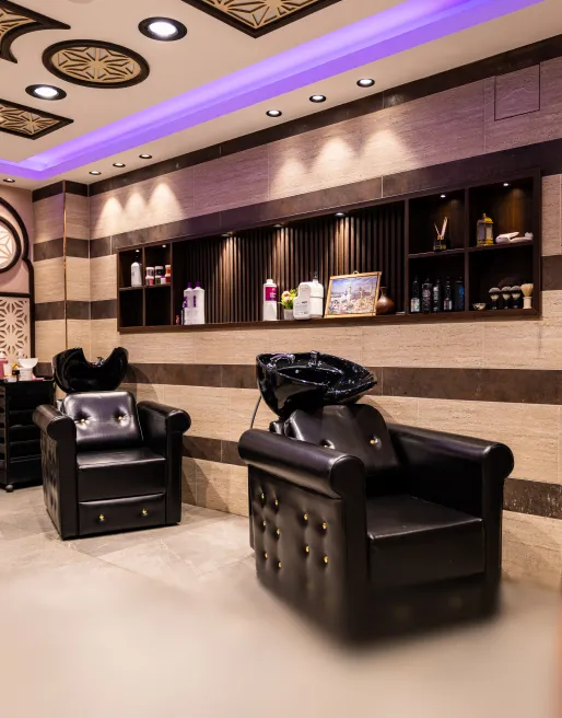 Moderner Friseursalon mit zwei schwarzen Ledersesseln und Waschstationen vor einer Wand mit Regalen und LED-Beleuchtung.