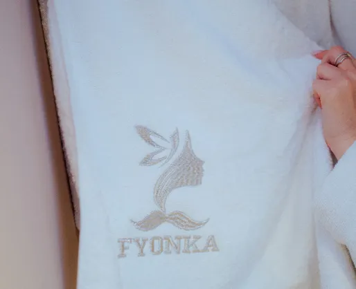 Weißer Bademantel mit einem Logo einer stilisierten Frauenprofil-Silhouette und dem Schriftzug 'FYONKA'.