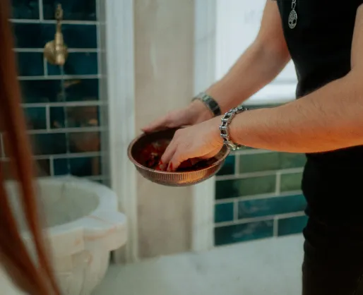 Person wäscht Hände mit rotem Wasser in einer traditionellen schwarzen Schüssel neben gefliester Wand.