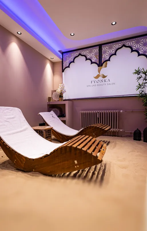 Entspannungsraum mit zwei ergonomischen Holzliegen auf Sandboden und einem beleuchteten Spa-Logo an der Wand.